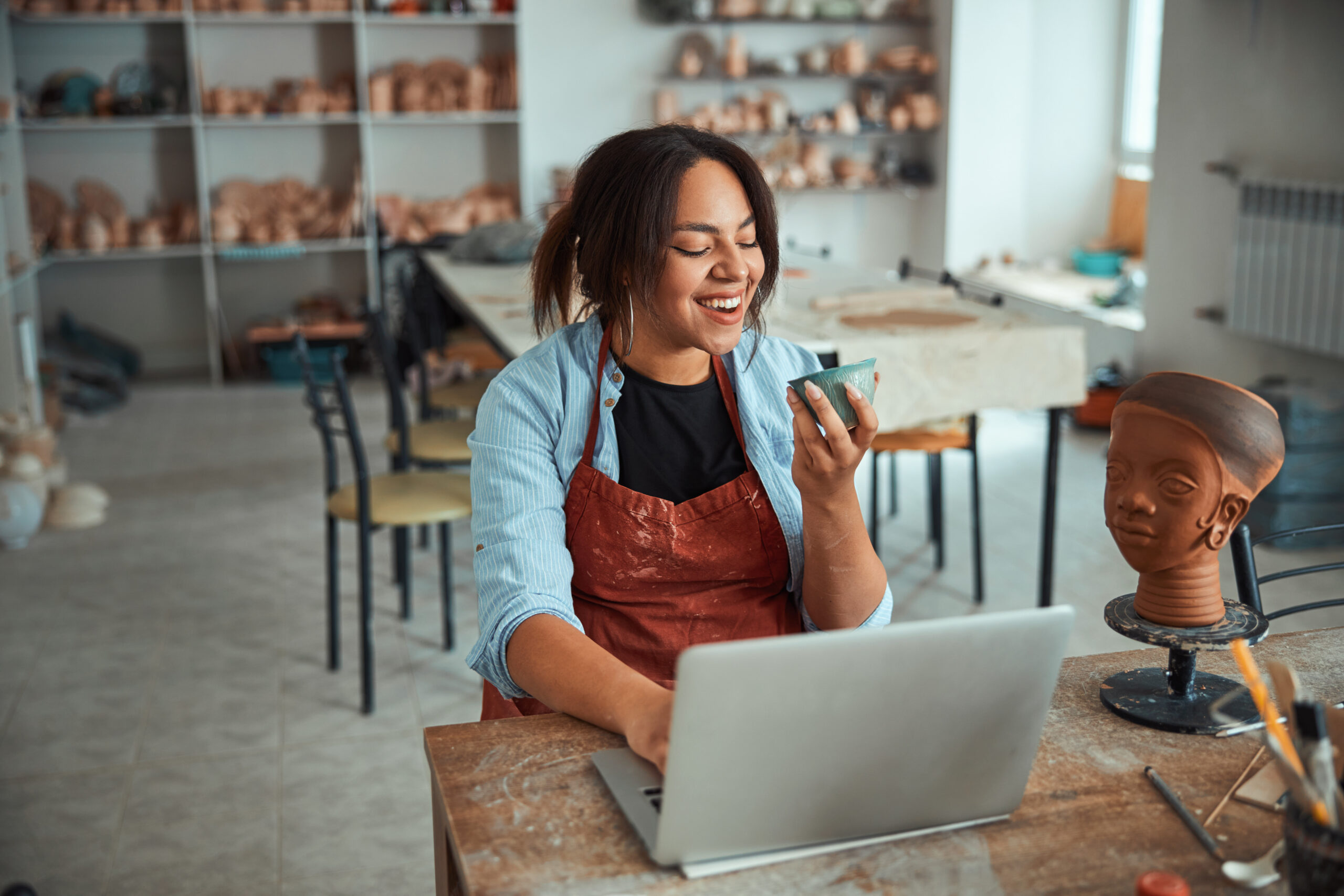 Building her online business presence: A woman wearing an apron smiles while holding a pottery piece in one hand and using a laptop with the other, in a studio filled with ceramics and sculptures. She's simultaneously enjoying her craft and building presence for her online business.