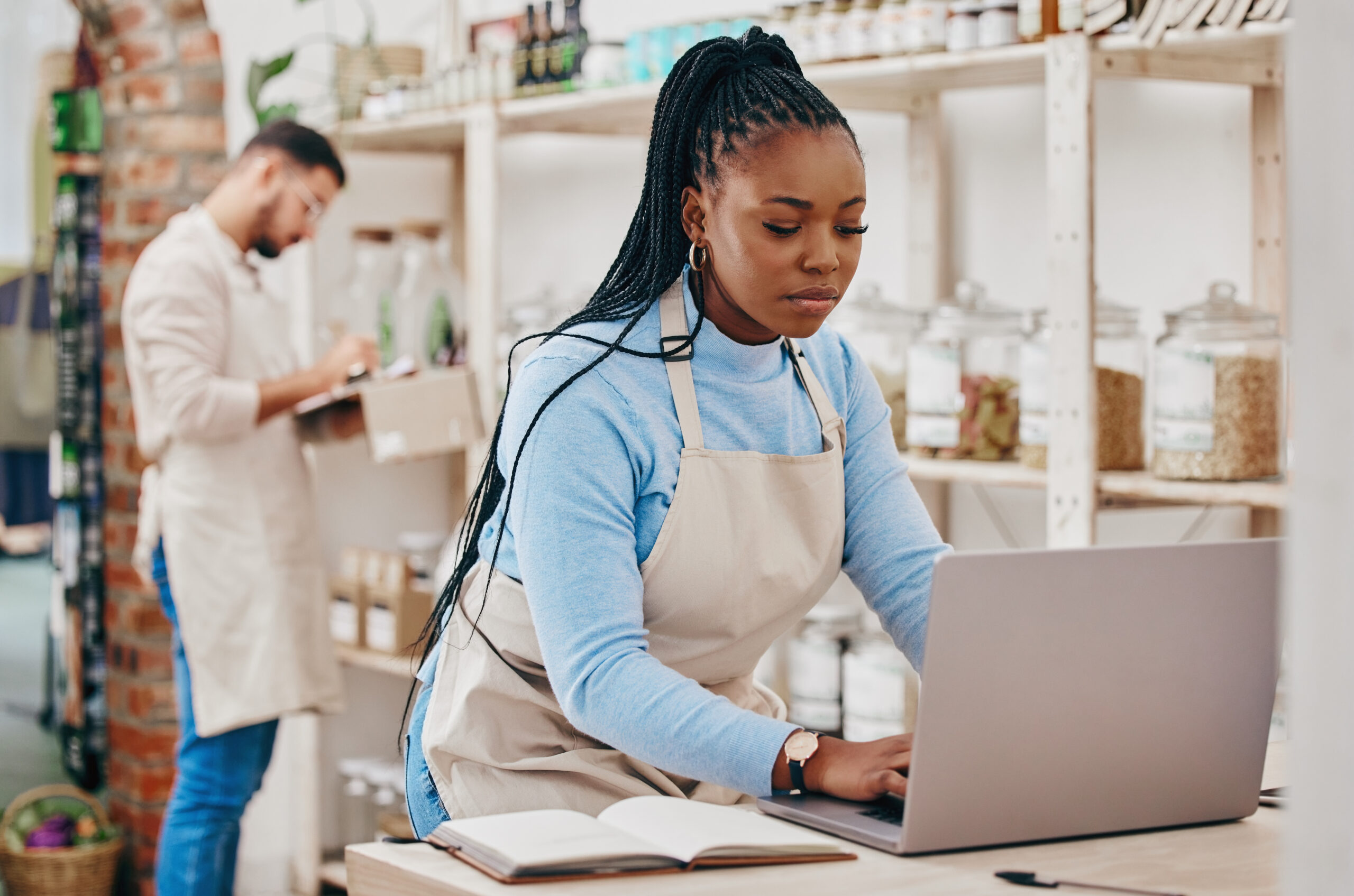 Two people in an indoor shop setting. One person, wearing a beige apron, types on a laptop with a notebook nearby, likely focusing on digital marketing strategies for small businesses. Another person in the background, also in a beige apron, writes on a clipboard.
