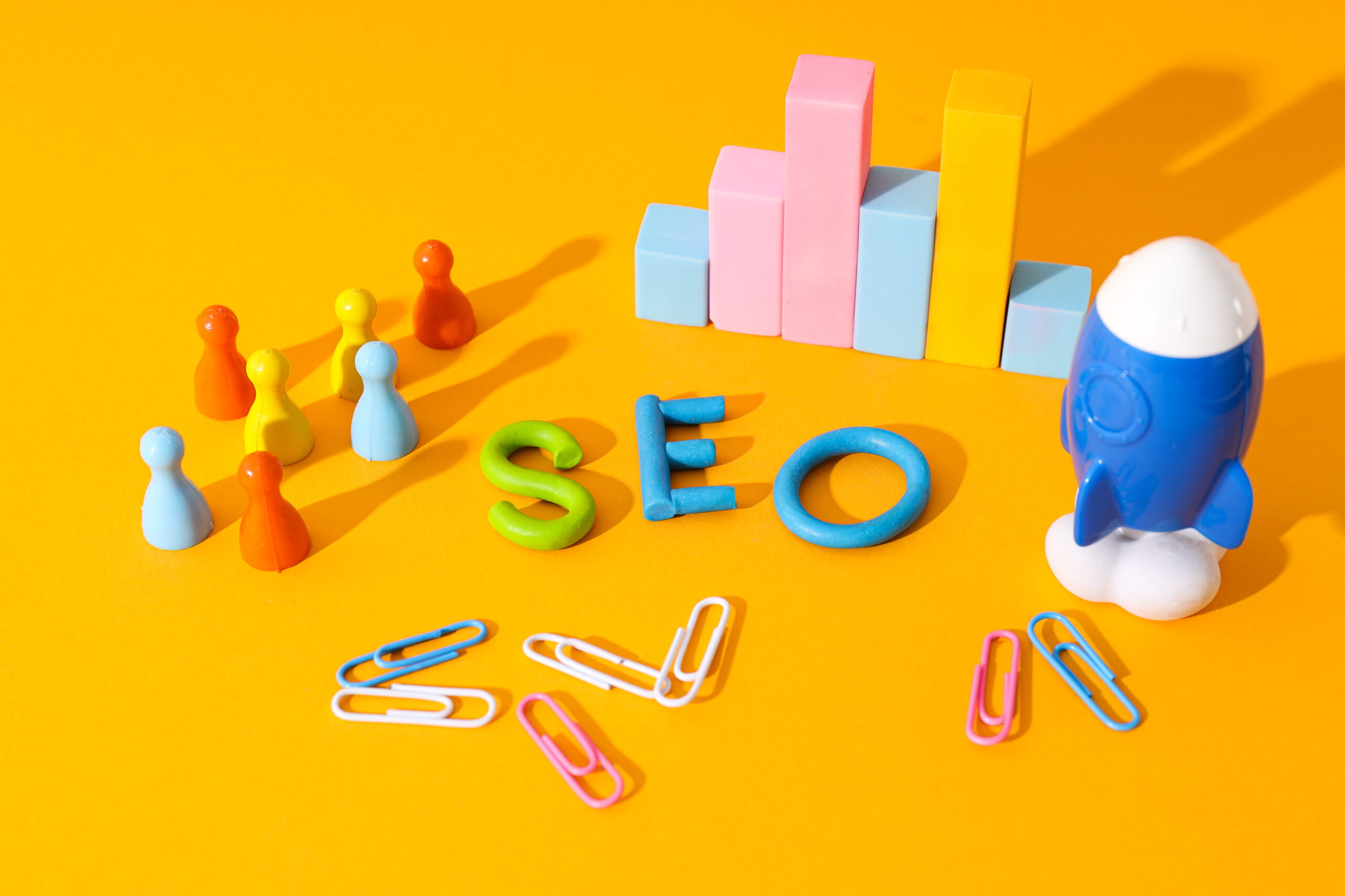 Colorful SEO letters, a group of multicolored game pieces, a blue toy rocket, and a graph made of blocks symbolize Local SEO strategies, all neatly arranged with paperclips on a bright yellow background.