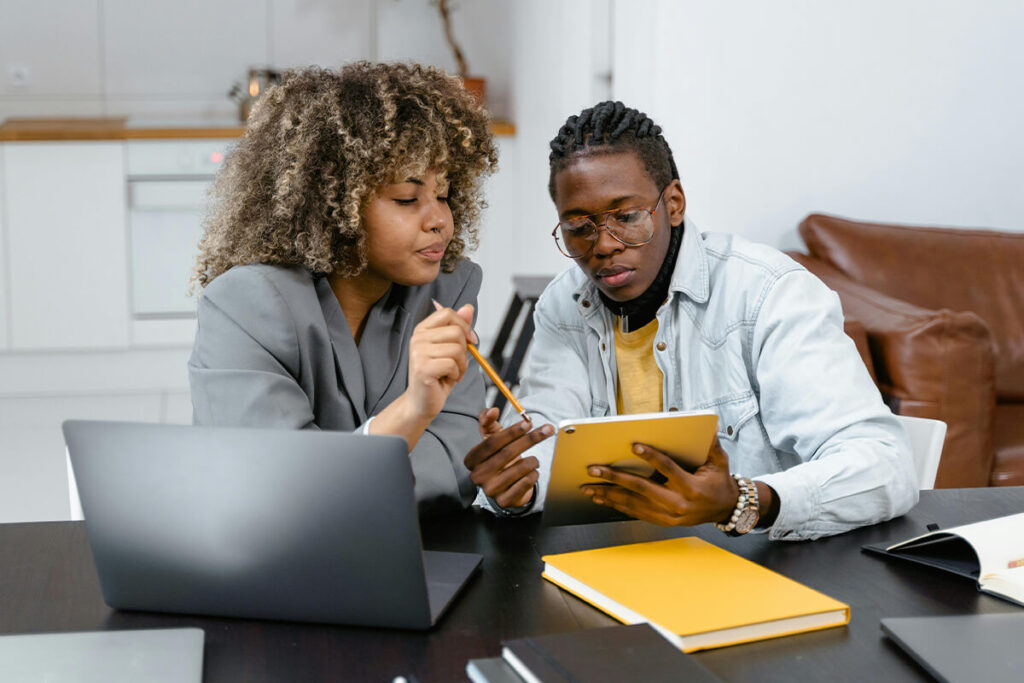 Two people sit at a table with a laptop and notebooks, discussing digital assets whilst looking at a tablet together, likely reviewing strategies for effective asset management. - Kemoso