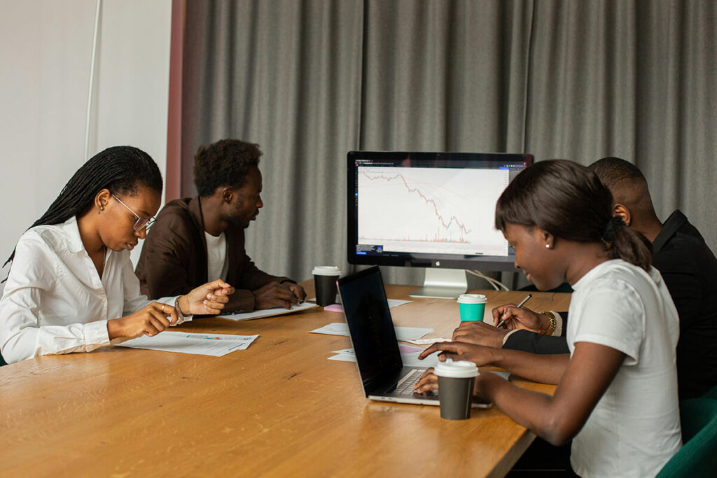 Four people sit at a table with laptops and papers, analysing data; a large monitor displays a declining line graph in the background, highlighting challenges in digital asset management. - Kemoso
