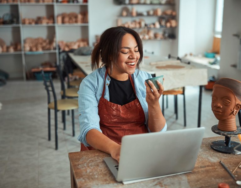 Building her online business presence: A woman wearing an apron smiles while holding a pottery piece in one hand and using a laptop with the other, in a studio filled with ceramics and sculptures. She's simultaneously enjoying her craft and building presence for her online business.