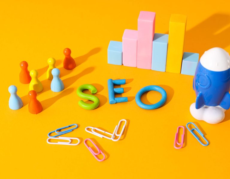 Colorful SEO letters, a group of multicolored game pieces, a blue toy rocket, and a graph made of blocks symbolize Local SEO strategies, all neatly arranged with paperclips on a bright yellow background.