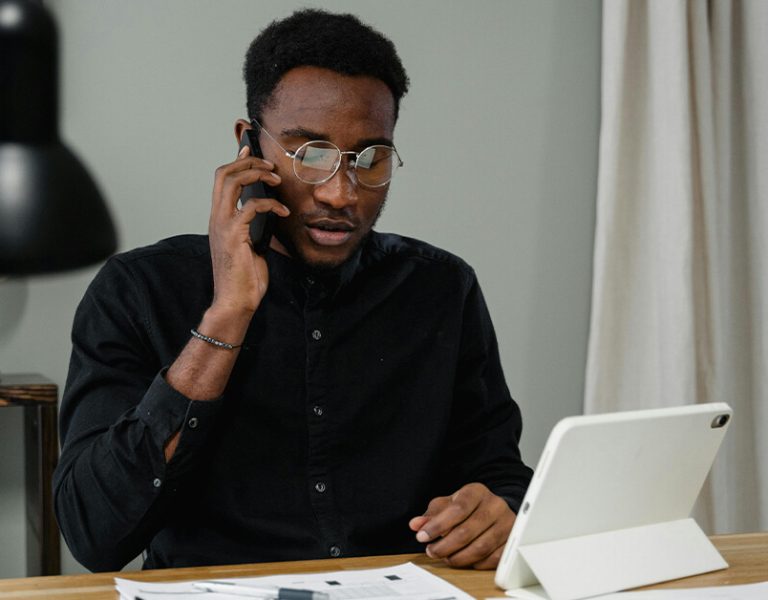 A man wearing glasses sits at a desk, talking on a mobile phone and looking at a tablet, managing Digital Assets Control with papers and a lamp nearby. - Kemoso