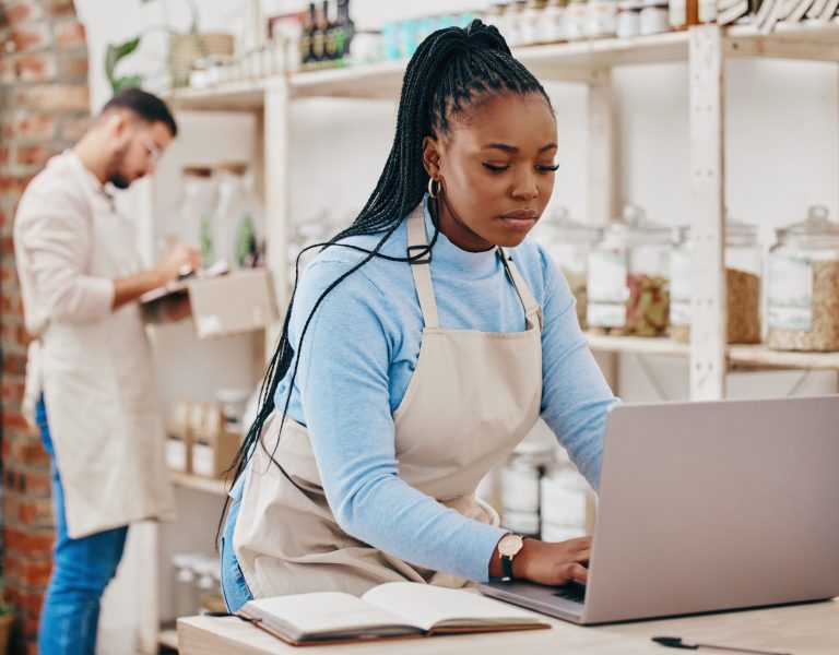 Two people in an indoor shop setting. One person, wearing a beige apron, types on a laptop with a notebook nearby, likely focusing on digital marketing strategies for small businesses. Another person in the background, also in a beige apron, writes on a clipboard.